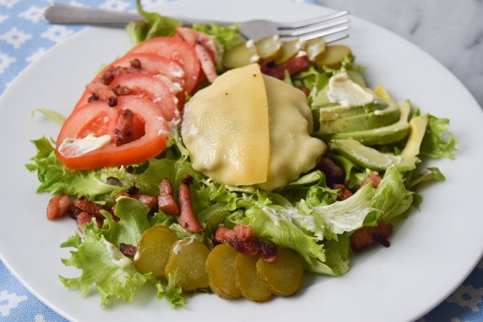 Een bord met een salade met een burger, gesmolten kaas, tomaat, avocado, spekjes en augurk
