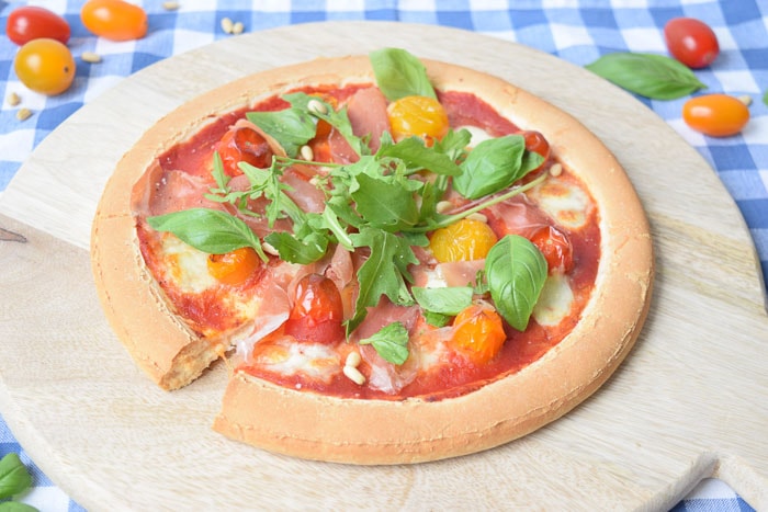 Een low FODMAP pizza parmaham op een houten plank met basilicum en tomaatjes ernaast