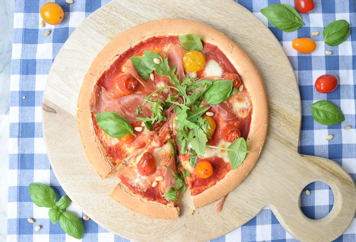 A low FODMAP pizza parmaham photographed from above with fresh basil and tomatoes next to it