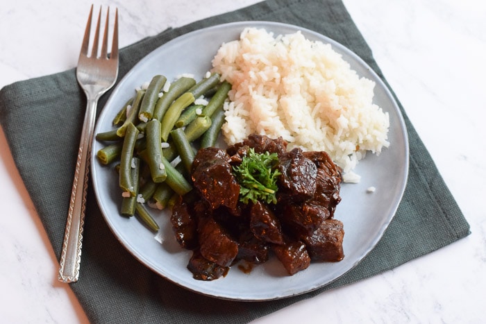 A low FODMAP beef stew on a plate with rice and green beans next to it. Underneath the plate is a green napkin and a fork next to it