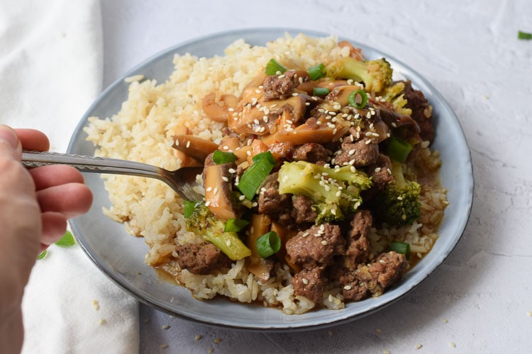 A plate with a mixture of beef and broccoli in soy sauce with rice and somebody taking a bit out of it