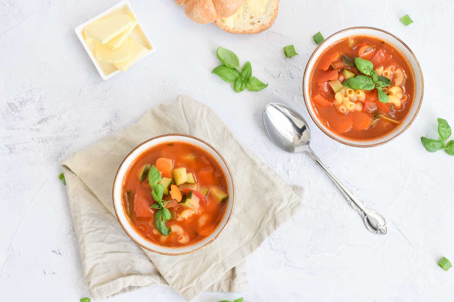 Two bowls of minestrone soup with a spoon in between and some bread and butter on the side