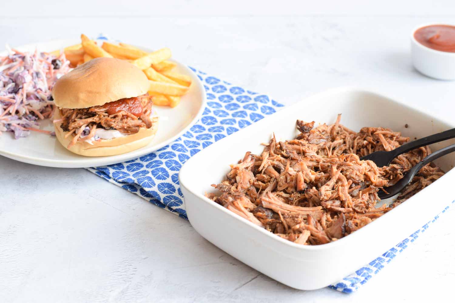 A bowl of pulled pork with a plate behind it with pulled pork on a bun, coleslaw and fries