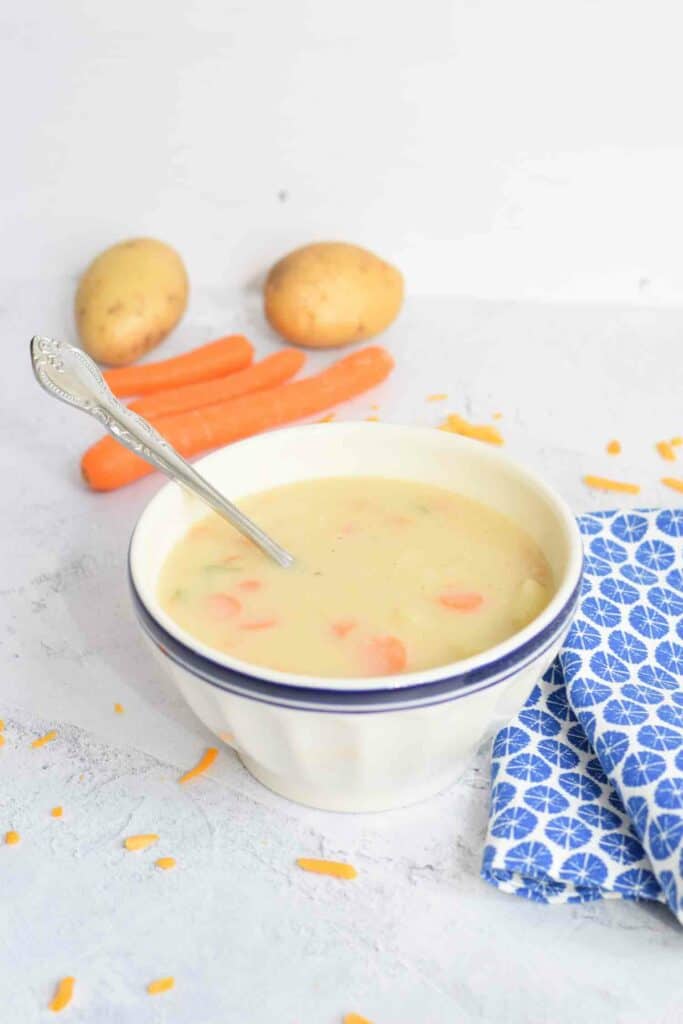 A bowl of low FODMAP potato soup with a spoon in it with on the background some potatoes and carrots