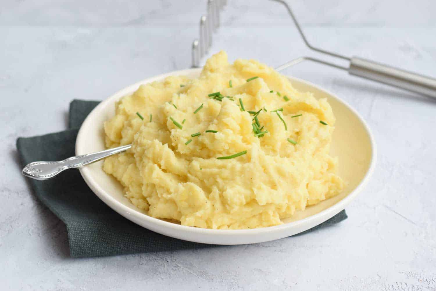 Low FODMAP mashed potatoes on a plate with a potato masher behind it