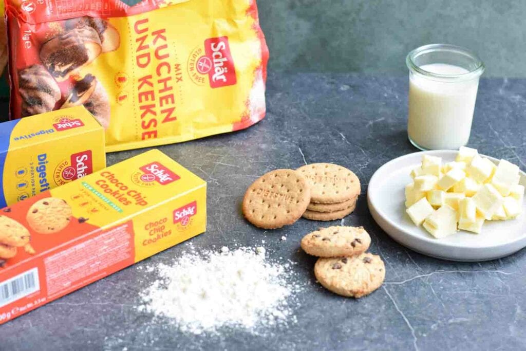 Een verpakking van Schär meelmix, digestives en choco chip koekjes met daarnaast wat koekjes, boter, melk en glutenvrij bloem