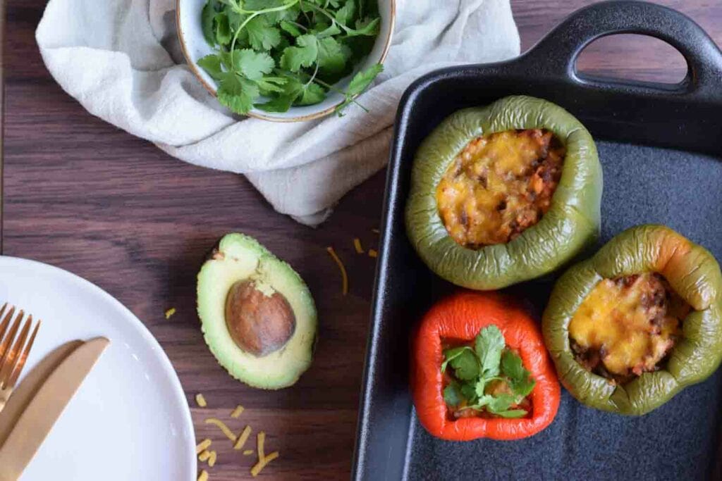 Drie low FODMAP gevulde paprika's in een ovenschaal met avocado, kaas en koriander ernaast